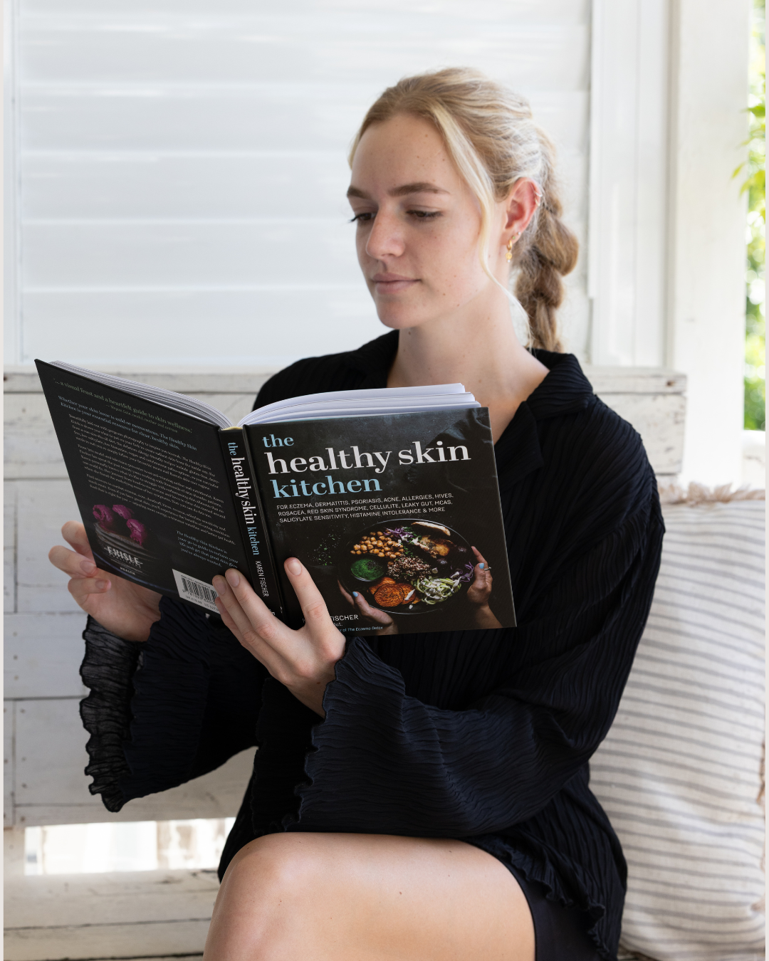 Woman reading 'The Healthy Skin Kitchen' book in a bright room.
